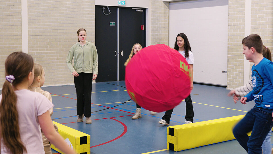 Kinderen gooien een grote roze bal naar elkaar over in een gymzaal.