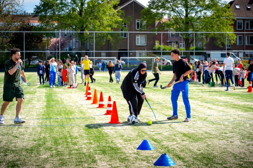 Een groep kinderen staat in een rij op een sportveld terwijl een volwassene met opgestoken armen hen lijkt aan te moedigen of instructies geeft.